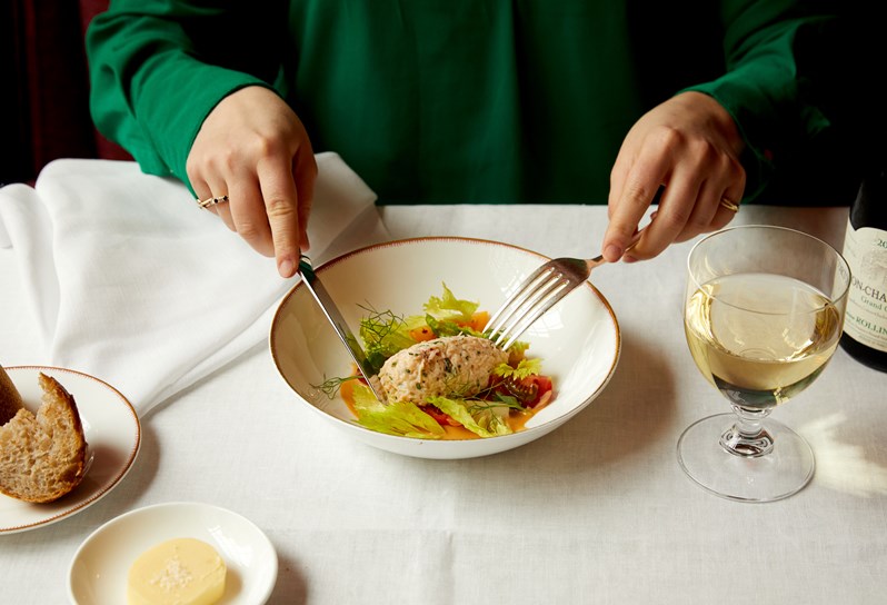 The bottom half of woman sat at a dining table. She is cutting into a dish of crab salad, there is a glass of white wine to the right and a piece of bread and butter dish to the left.