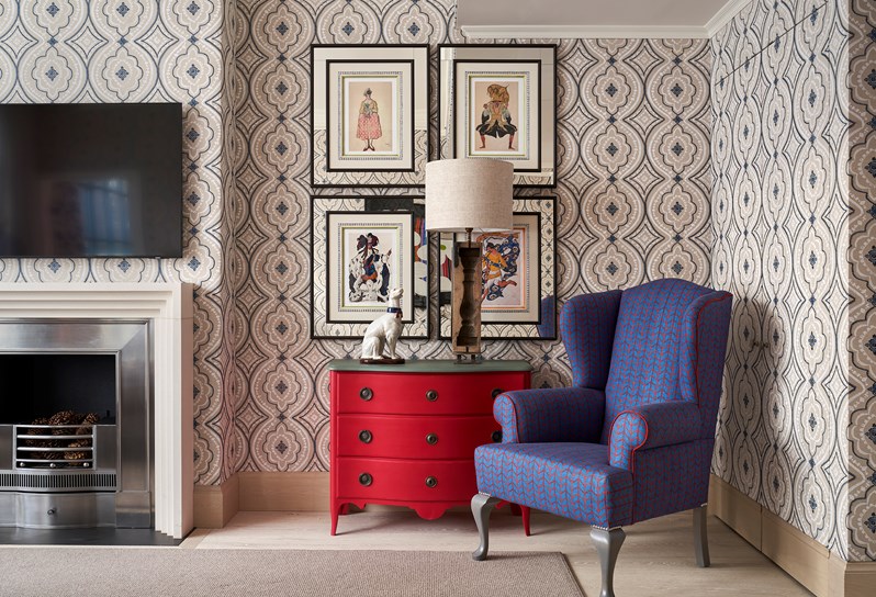 2 framed figure paintings on a white and blue patterned wall. In front is a red chest of drawers holding a white dog statue and a white lamp. A blue and red patterned Wing chair to the left.