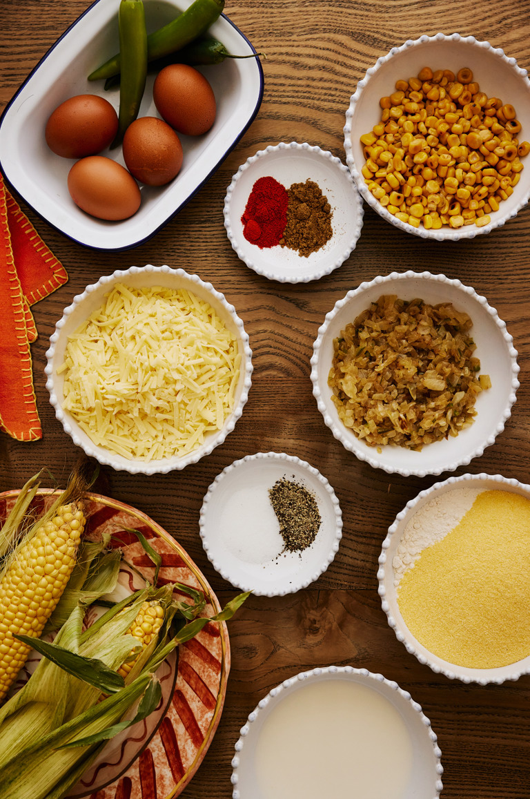 In this image there is multiple white dishes with ingredients used to make the cornbread. In the bottom right, you can see a Sasha Compton filled with fresh corn on the cob, in the white dishes you can see milk, corn flower, baking flower, softened onions, grated cheese, corn and seasonings and in a white and blue baking dish in the top left corner chillis and eggs.
