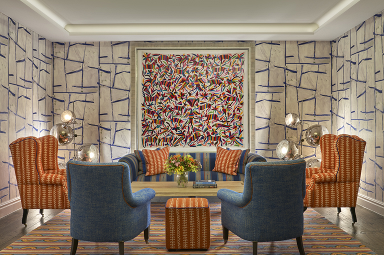 An image of the seating area in the Sandra Blow private events room at The Soho Hotel. The image shows a sofa in a blue and orange colour scheme under a piece of art, with two orange armchairs flanking the sofa and two blue arm chairs at the front facing away from the camera. 