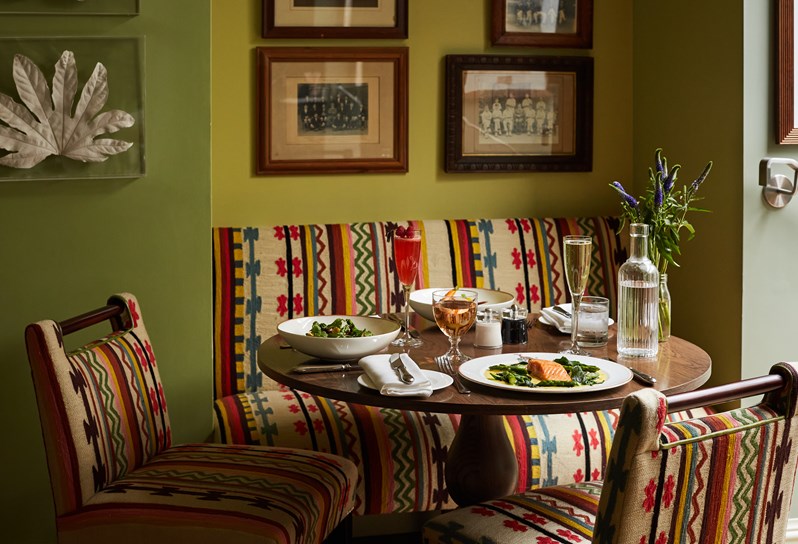 A round wooden dining table display, a plate of salmon with vegetables, and two bowls of salad with cutlery, napkins and side plates. There is also a glass of champagne, wine, a pink cocktail and a bottle of water with a small vase of flowers. There are two chairs and banquette seating in a multicoloured geometric fabric.