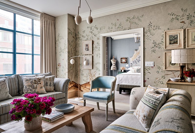 A neutral coloured floral patterned sofa, cushions and wallpaper opposite a blue striped sofa, a wooden coffee table in the middle. A bed and mannequin can be seen through an open door.