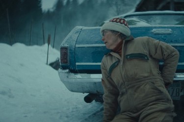 A still from 'Dead of Winter'. Against a snowy backdrop, an older woman dressed in a snowsuit and beanie hat crouches behind a car, turning turning her head as if to look around the car.