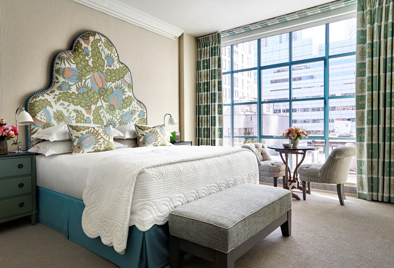 A bed with a headboard upholstered in a pale blue, green and pink floral pattern on a white background and a grey ottoman at the end. Two striped blue chairs and a coffee table in front of the window which have curtains in Ozone fabric in a blue colour scheme.