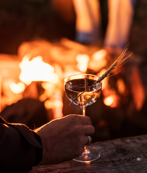 A hand holding a cocktail by a firepit