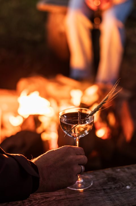 A hand holding a cocktail by a firepit