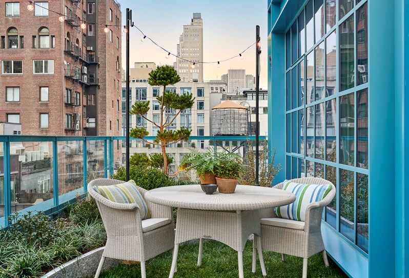 A bedroom terrace during the day. A woven outdoor dining table and two chairs, surrounded by plants and with grass underneath. High rise buildings can be seen in the background.