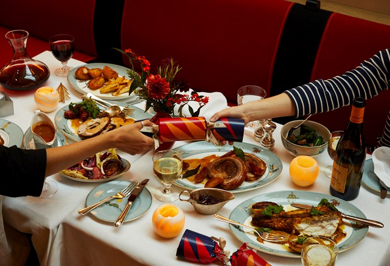 Festive table with numerous different festive dishes on Sailor's Farewell crockery, some glasses on wine and two people on either side of the table both holding a Christmas cracker