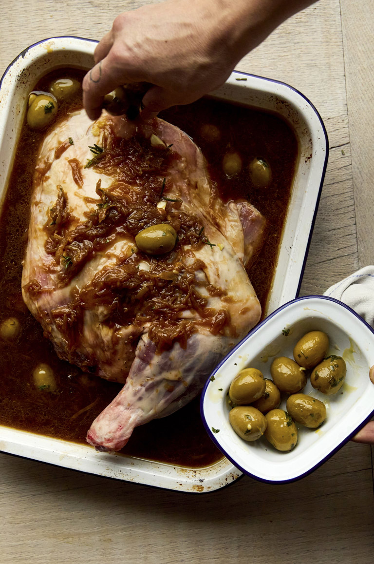 Portrait shot of a lamb joint, with a chef's hand placing olives around it