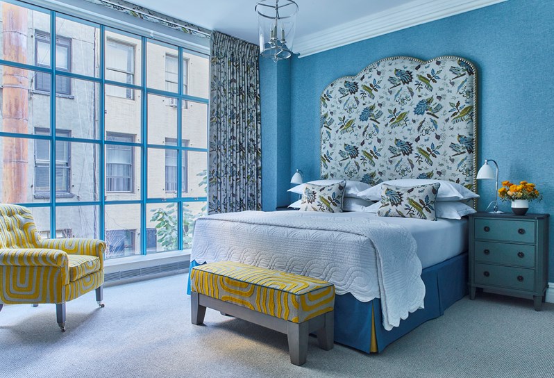 A bed with a blue and yellow patterned headboard and cushions and matching curtains. A matching yellow patterned ottoman and armchair.