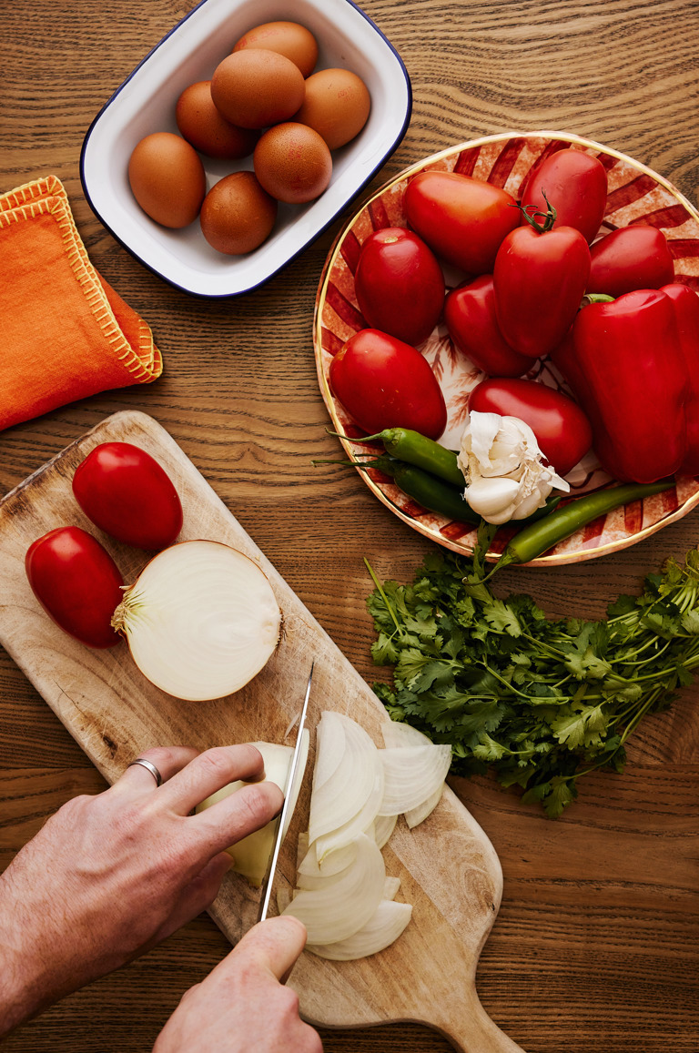 In this image you can see hands chopping onions on a wooden board prepping vegetables to make the sauce base of the baked eggs. There is also a fresh bunch of coriander on the right hand side and Sasha Compton of fresh tomatoes, peppers, garlic and chillis.