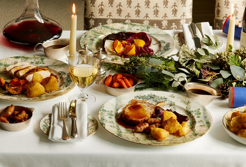 A closeup of a table setup for festive dining. There are Christmas crackers, candles, and plates with mains being laid out, alongside a glass of white wine and a bottle of red wine in the decanter. The mains include all sorts of festive roasts, such as potatoes, pigs in a blanket, carrots, chicken, pork and gravy. The crockery being used is Tall Trees.