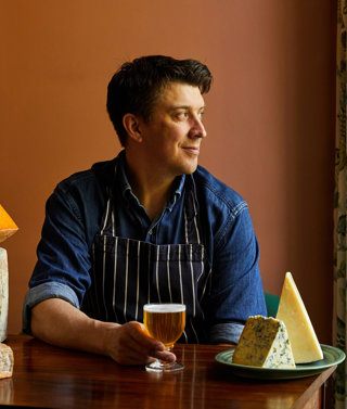 An image of Joe Fox holding a glass of beer surrounded by different cheeses at The Potting Shed, Dorset Square Hotel. 