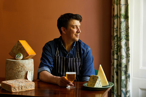 An image of Joe Fox holding a glass of beer surrounded by different cheeses at The Potting Shed, Dorset Square Hotel. 