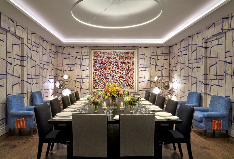 An image of the long dining table set up with flowers, place mats, plates, cutlery and glassware ready for dining in the Sandra Blow, private events room, at The Soho Hotel. With 14 grey chairs around the long tables.