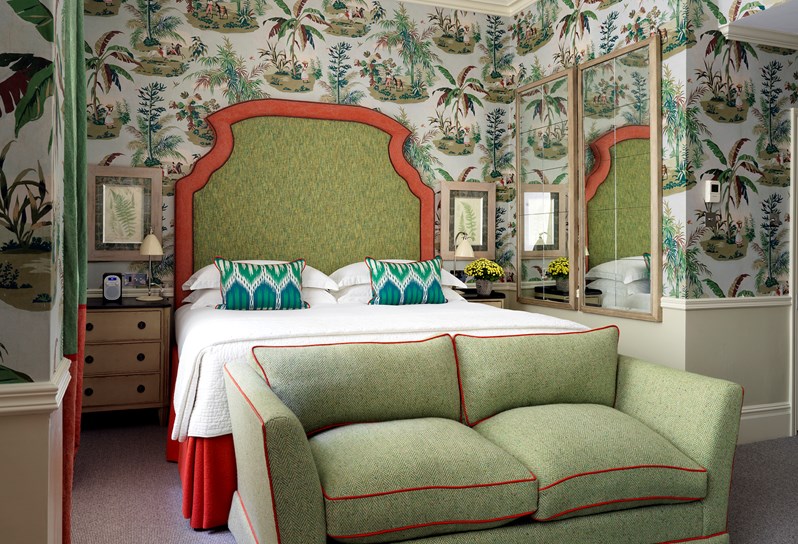 An angled bed shot in Room 111, at Covent Garden Hotel. There is a large green and orange headboard, curtains, sofa as well as a tropical themed wallpaper.