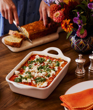 This images a portrait image that shows the final product of the recipe of Baked eggs and cornbread, here you can see a white baking dish with baked eggs in a totamto sauce topped with feta and corriander. in the bakground you can see a lady in a blue dress cutting into cornbread on a wooden board. in the foreground there is a stack of mythical creatures side plates and an orange napkin from Kit Kemp shop.