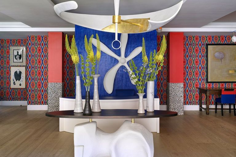  A wide shot of the lobby reception area featuring a central table and a large sculptural light hanging above. In the background is a reception desk with a large sculptural flower hung on the blue wall behind. The lobby area is bright and colourful with patterned fabric adorning the walls and statement pieces of artwork at 9-11 Richmond Buildings