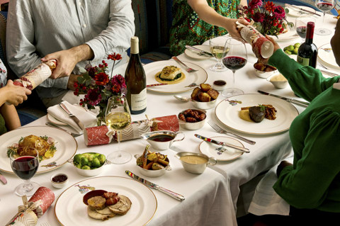 A table of six set up for Christmas dining at Brumus. There are plates of meat and fish with small side dishes and bottles of wine. Four people can be seen partially in view pulling crackers across the table