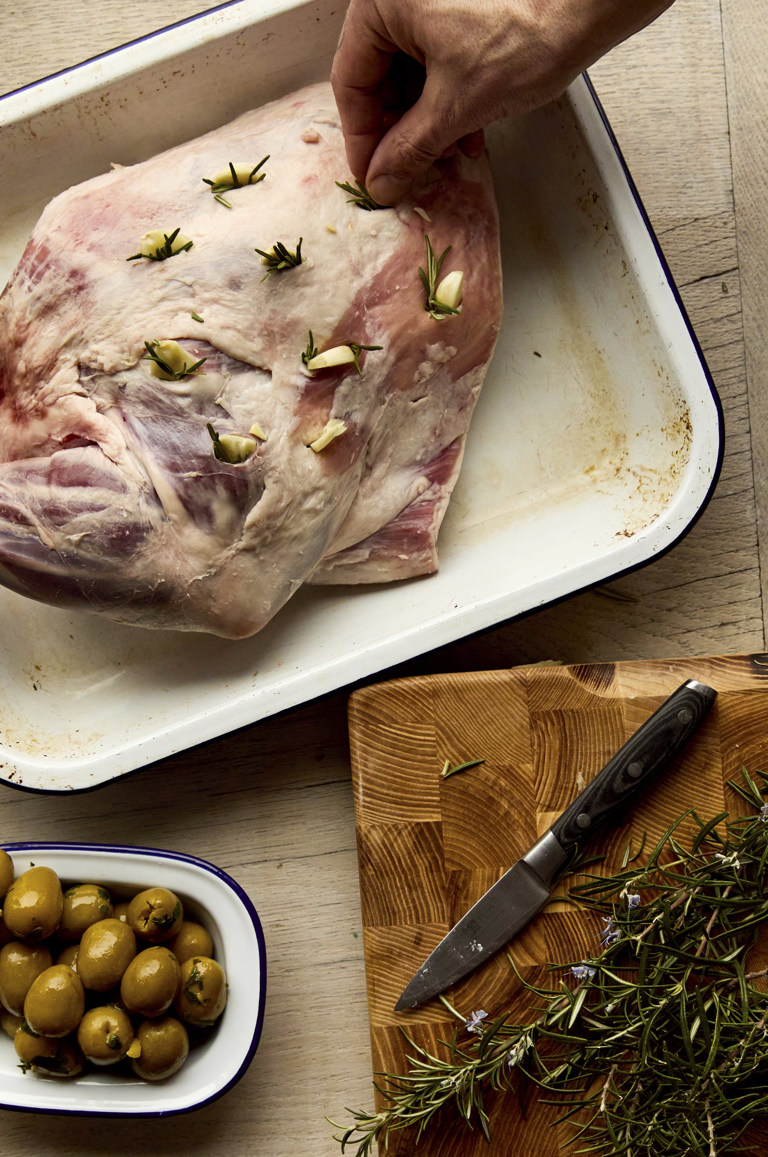A shot from above of a raw lamb joint with pieces of garlic being placed in the meat. Next to it, there is a bowl of olives and herbs on a chopping board