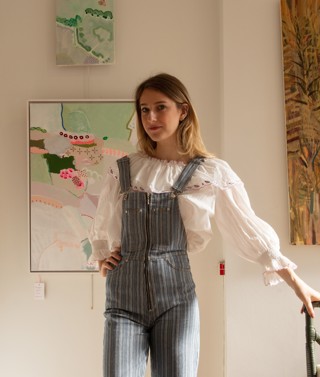 Artist and curator Domenica Marland dressed in dungarees, leaning on a small chair. Her framed paintings hang on white walls behind her