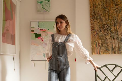 Artist and curator Domenica Marland dressed in dungarees, leaning on a small chair. Her framed paintings hang on white walls behind her