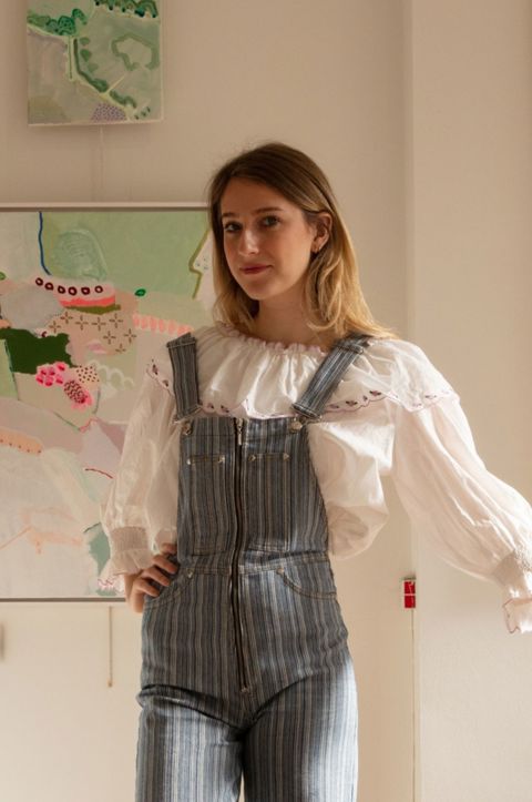 Artist and curator Domenica Marland dressed in dungarees, leaning on a small chair. Her framed paintings hang on white walls behind her