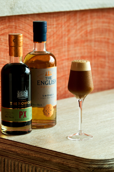 An image of a brown, smoky cocktail, frothy on top. Next to a bottle the The English Smokey whiskey and The Norfolk PX.