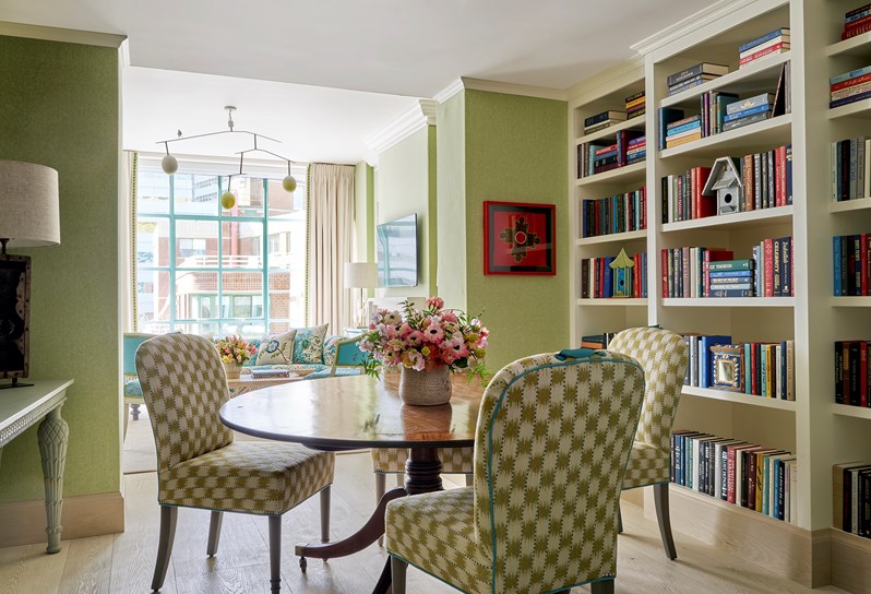 A room with green walls and a large bookshelf on the right side. A round dining table with pink and green flowers on, surrounded by four Susan chairs in a green and white checkered fabric. A blue sofa with floral patterned cushions can be seen in front of windows in the next room.