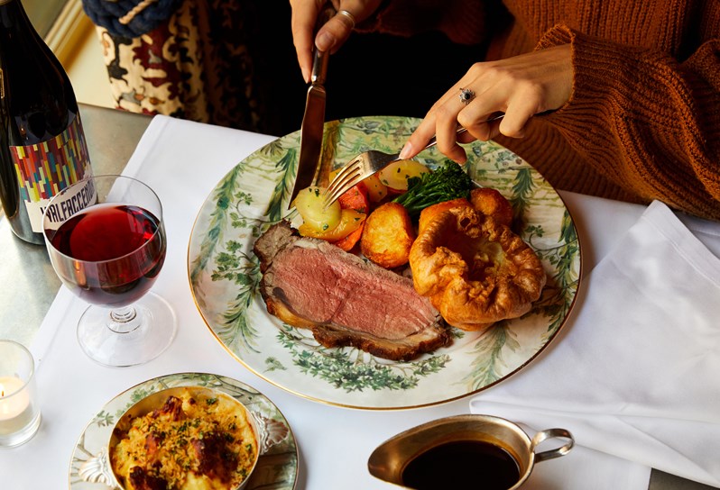 Sunday roast on tall trees crockery with cutlery held by a woman. A glass of red wine and jug of gravy on the side
