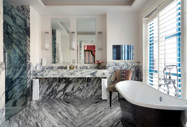 A black and white marble tiled bathroom. A walk in shower to the left and a black and white bathtub in front of windows with white shutters to the right. On the back wall, two mirrors above a marble surface with two sinks and towels hanging either side.