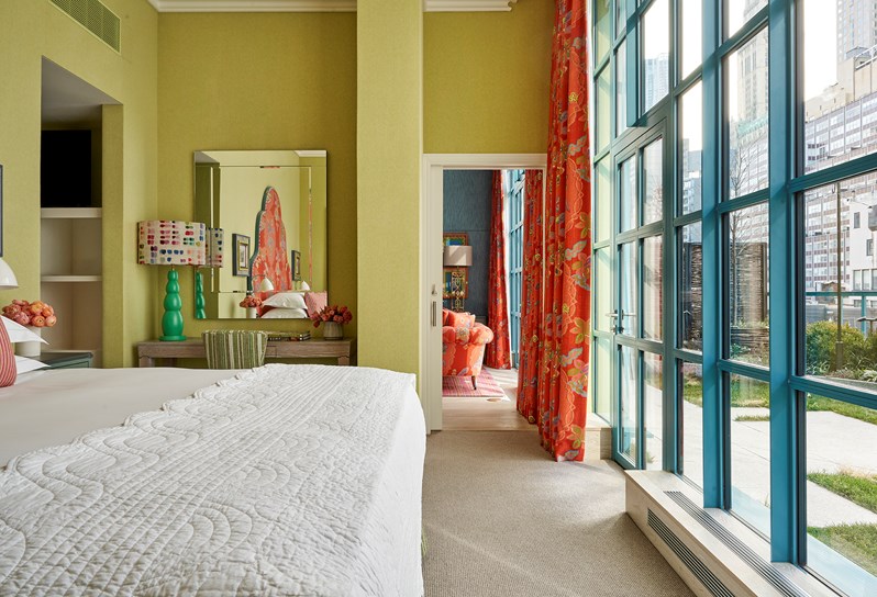 A green walled room. A bed is partially in view on the left hand side, the orange headboard can be seen in the reflection of the mirror on the wall. There is a green striped chair and a desk with a turquoise lamp on underneath the mirror. To the right, there are floor to ceiling windows with curtains in orange 'Chainstitch' fabric to match the headboard.