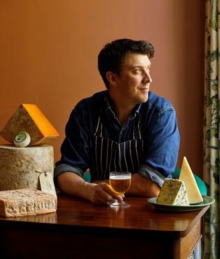 An image of Joe Fox holding a glass of beer surrounded by different cheeses at The Potting Shed, Dorset Square Hotel. 