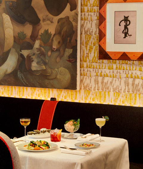 An image to promote the Summer à la carte starters at Ham Yard Bar & Restaurant at Ham Yard Hotel. The image shows a white-linen table set with a selection of starters, including oysters, prawn cocktail, and vibrant salads. Three cocktails are placed alongside the dishes, set against a backdrop of patterned wallpaper and framed artwork