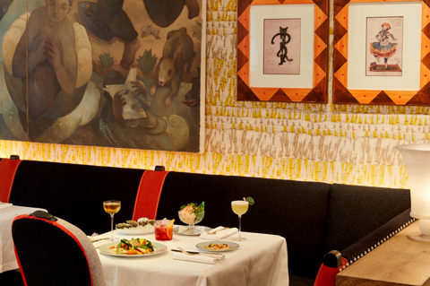 An image to promote the Summer à la carte starters at Ham Yard Bar & Restaurant at Ham Yard Hotel. The image shows a white-linen table set with a selection of starters, including oysters, prawn cocktail, and vibrant salads. Three cocktails are placed alongside the dishes, set against a backdrop of patterned wallpaper and framed artwork