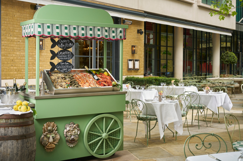 Wide shot of the exterior of Ham Yard Hotel featuring a green oyster cart and tables set up for outdoor dining