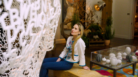 Shot of design director Willow Kemp, sitting on a wooden bench looking up at pink lettered art piece. 