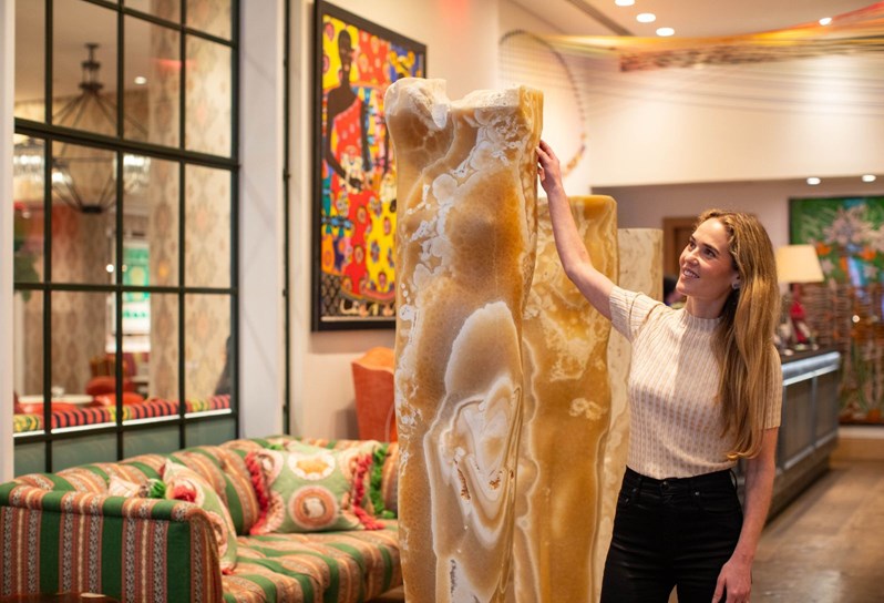Firmdale Hotels Art Ambassador and Design Director, Willow Kemp stands in the lobby of The Whitby Hotel. She is reaching her right arm up to touch a large beige stone sculpture, with two smaller sculptures behind her. The lobby's sofa area and reception desk can be seen out of focus behind her.