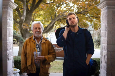 A still from the film 'Tuner': the image portrays two men waiting at a doorstep, to the left the man is wearing a yellow jacket and to the right the man wears a navy shirt. 