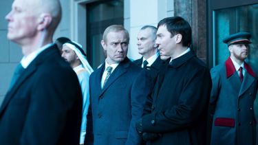 A still from the film "The Wizard of the Kremlin": A group of men are stood outside a building and one man wearing a navy blazer looks over at the man next to him
