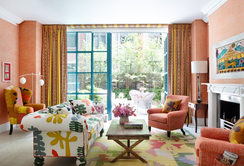 A living room space with orange walls, a multicoloured sofa and orange chairs. Bifold doors open onto a terrace with a table and chairs