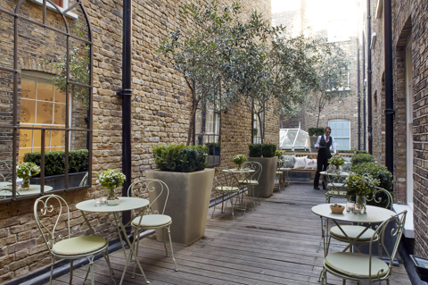 A narrow terrace lined with tables of two on the first floor at Haymarket Hotel. A waitress holds a tray at the far end