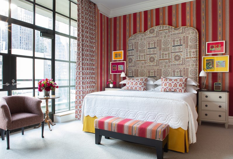 A bright and colorful bedroom in tones of yellow, red and orange, containing a king sized bed with a large headboard, a end of bed ottoman and a comfortable arm chair and bespoke wooden coffee table with fresh flowers in front of the floor to ceiling windows.