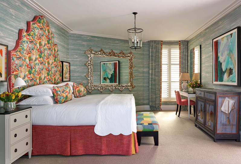 A side on image of the bedroom of The Deluxe Two Bedroom Suite, Room 5. A stylish bedroom featuring a vibrant patterned headboard and pillows, blue textured wallpaper, eclectic artwork, and a arm chair in the room.