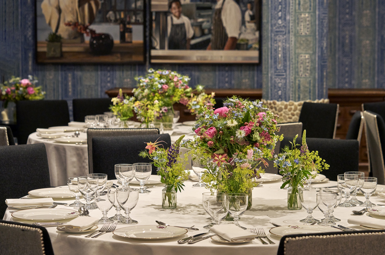 An close up image of the round dining table set up in the Indigo Room, there are white table cloths, floral bouquets in the centre and candles. each place around the table has a placemat, plate, cutlery, glassware and napkins ready for dining. private events room at The Soho Hotel.