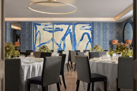 An image of multiple round dining table set up in the Indigo Room, there are white table cloths, floral bouquets in the centre and candles. each place around the table has a placemat, plate, cutlery, glassware and napkins ready for dining. private events room at The Soho Hotel.