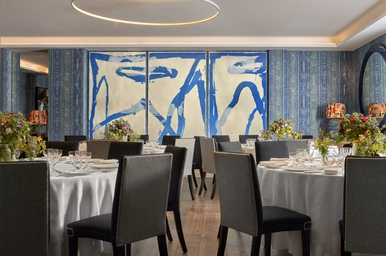 An image of multiple round dining table set up in the Indigo Room, there are white table cloths, floral bouquets in the centre and candles. each place around the table has a placemat, plate, cutlery, glassware and napkins ready for dining. private events room at The Soho Hotel.