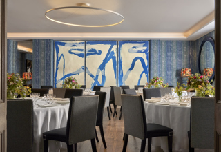 An image of multiple round dining table set up in the Indigo Room, there are white table cloths, floral bouquets in the centre and candles. each place around the table has a placemat, plate, cutlery, glassware and napkins ready for dining. private events room at The Soho Hotel.