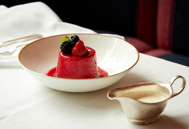 A small red berry sponge with mixed berries and coulis on top in a bowl. There is a small silver jug of cream to next to it.