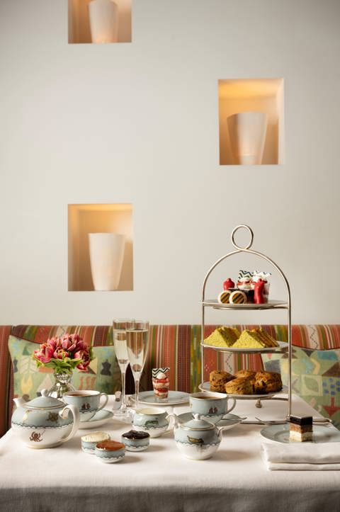 A portrait shot of the Kit Kemp Design Stories Afternoon Tea set up at The Whitby Bar. There is a cake stand with scones, cakes decorated with Kit Kemps Famous fabrics, edible mannequins and sandwiches with tea and Champagne